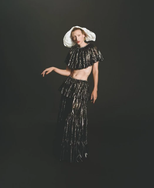 Elegant individual poses in a metallic fringe outfit and white hat on a dark backdrop in a studio fashion portrait, showcasing high fashion.
