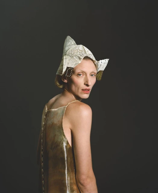Adult female with blonde hair wearing gold dress and paper origami hat looking over shoulder against dark grey studio backdrop.