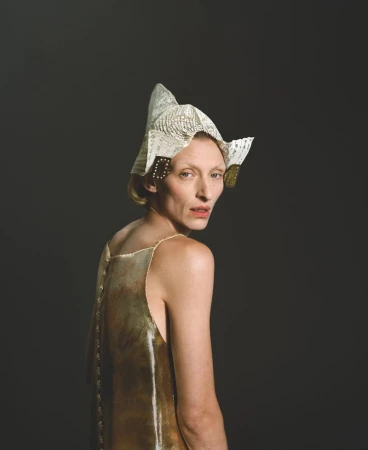 Adult female with blonde hair wearing gold dress and paper origami hat looking over shoulder against dark grey studio backdrop.