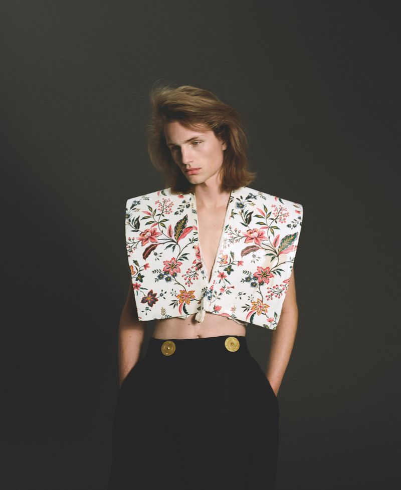 Young adult poses in a floral-patterned, cropped top with black high-waisted pants, hands in pockets, on a dark grey backdrop.