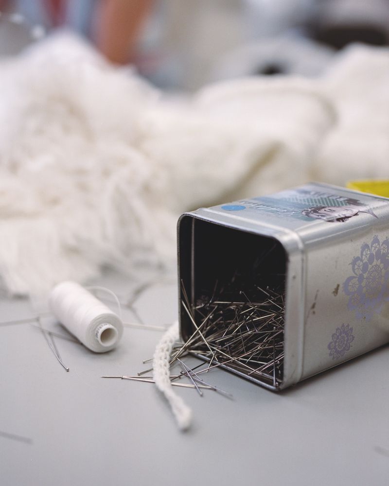 Close-up of sewing pins in a metal container with spool of white thread and lace, showcasing tailoring and crafting details.