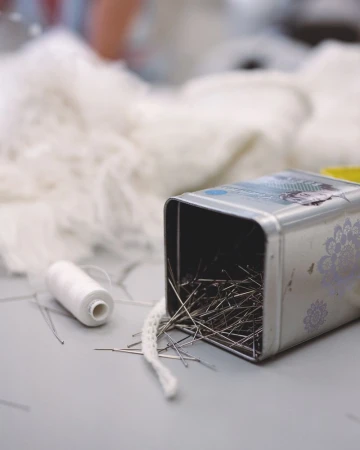 Close-up of sewing pins in a metal container with spool of white thread and lace, showcasing tailoring and crafting details.