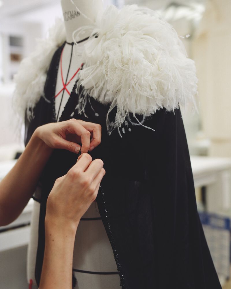 Close-up of a mannequin wearing a black garment with white feathered shoulders, adjusted by a person's hands in a bright room.