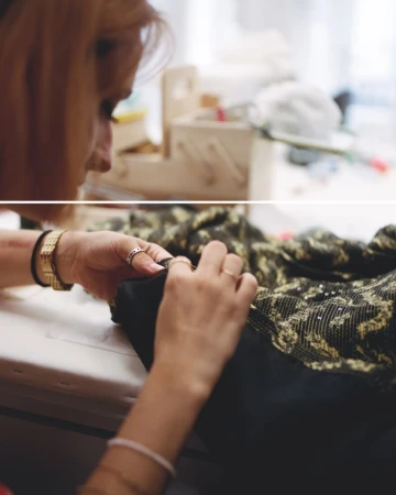 Woman sewing black fabric with gold detailing at a bright work table. Hands work with pins and rings visible. Indoor shot, focus on clothing detail.