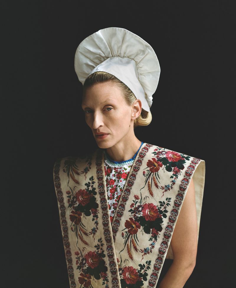 Portrait of adult woman wearing a chef's hat and floral shawl against a dark background in a studio photograph, fashion editorial style.