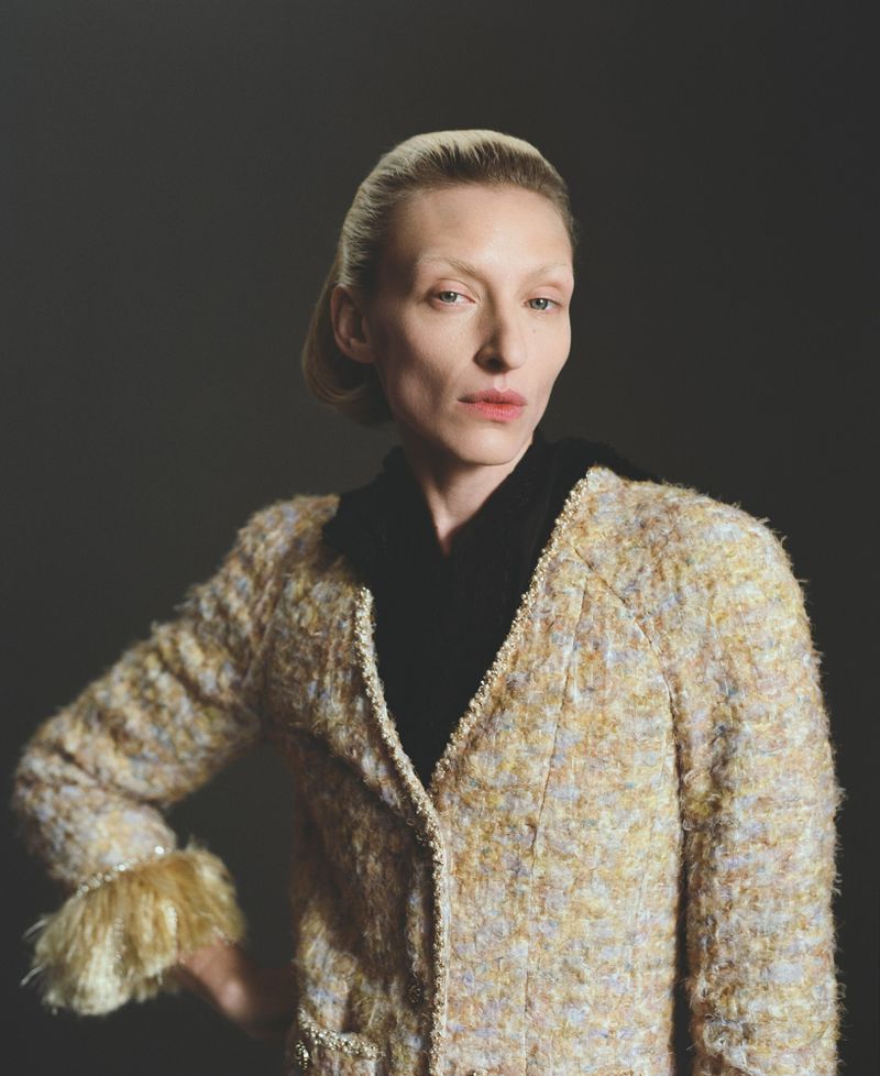 Portrait of an adult woman in a textured jacket, featuring fuzzy trim on the sleeves, against a solid dark gray studio backdrop.