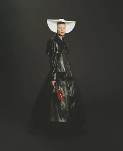 Maggie Maurer wearing a black leather coat and traditional white bonnet, holding a red flower against a dark background.
