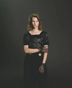 Model with shoulder-length hair wearing a black square-neck top, black trousers, and sheer lace gloves against a dark grey backdrop.