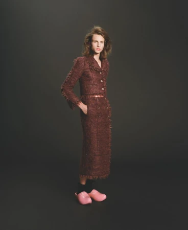 Model Maggie Maurer poses in a fringed brown tweed skirt suit and pink clogs against a dark studio background.