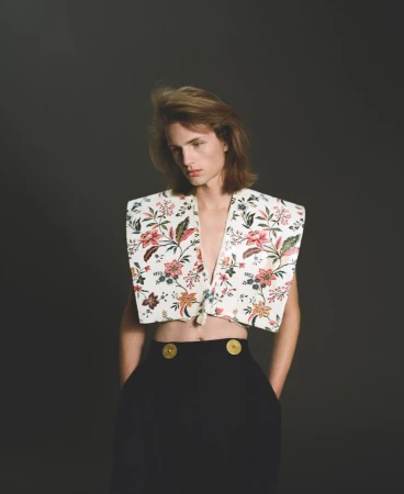 Model wearing a structured floral cropped vest and high-waisted black trousers with gold buttons against a dark backdrop.