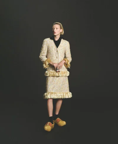 Model Maggie Maurer poses in a fringed cream tweed skirt suit and yellow painted wooden clogs against a dark background.