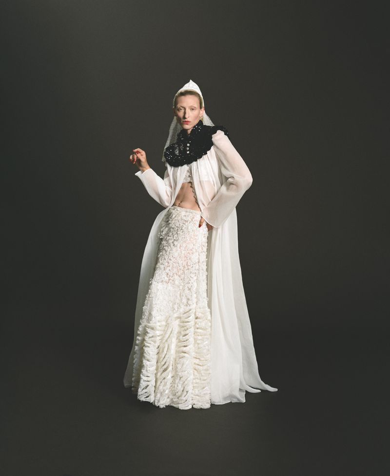Model Maggie Maurer poses in a sheer white hooded cape, textured maxi skirt, and black ruffled collar against a dark studio background.