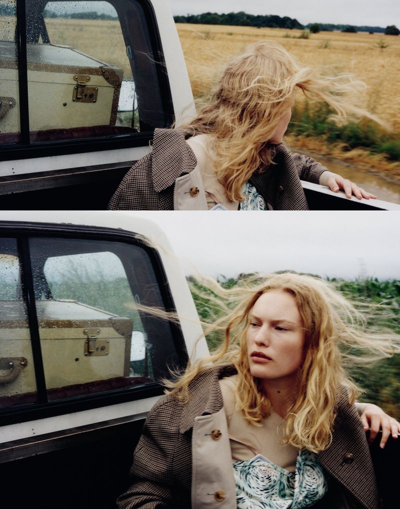 Woman rides in the back of a pickup truck with luggage. The scenery shows an open field under an overcast sky with windblown hair.