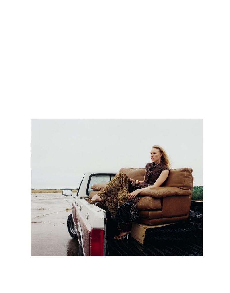 Fashion shot of a blond adult female model sitting on a brown leather sofa in the truck bed of a vintage pick-up in a field.