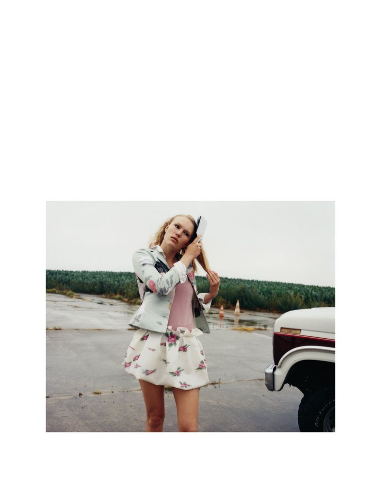 Young woman with fair hair wearing a floral skirt brushing her hair in an open field beside a classic white and maroon truck.
