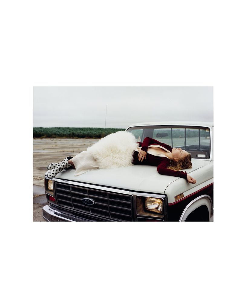 A woman wearing a cream-colored outfit and polka dot shoes lies on the hood of a white pickup truck in a rural outdoor setting.