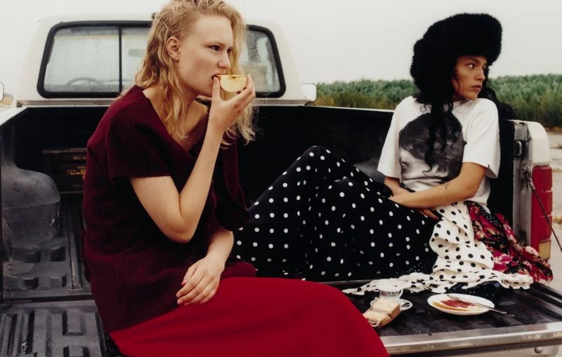 Two women sit in the back of a pickup truck, one eating an apple and the other with a furry hat, in a rural setting.