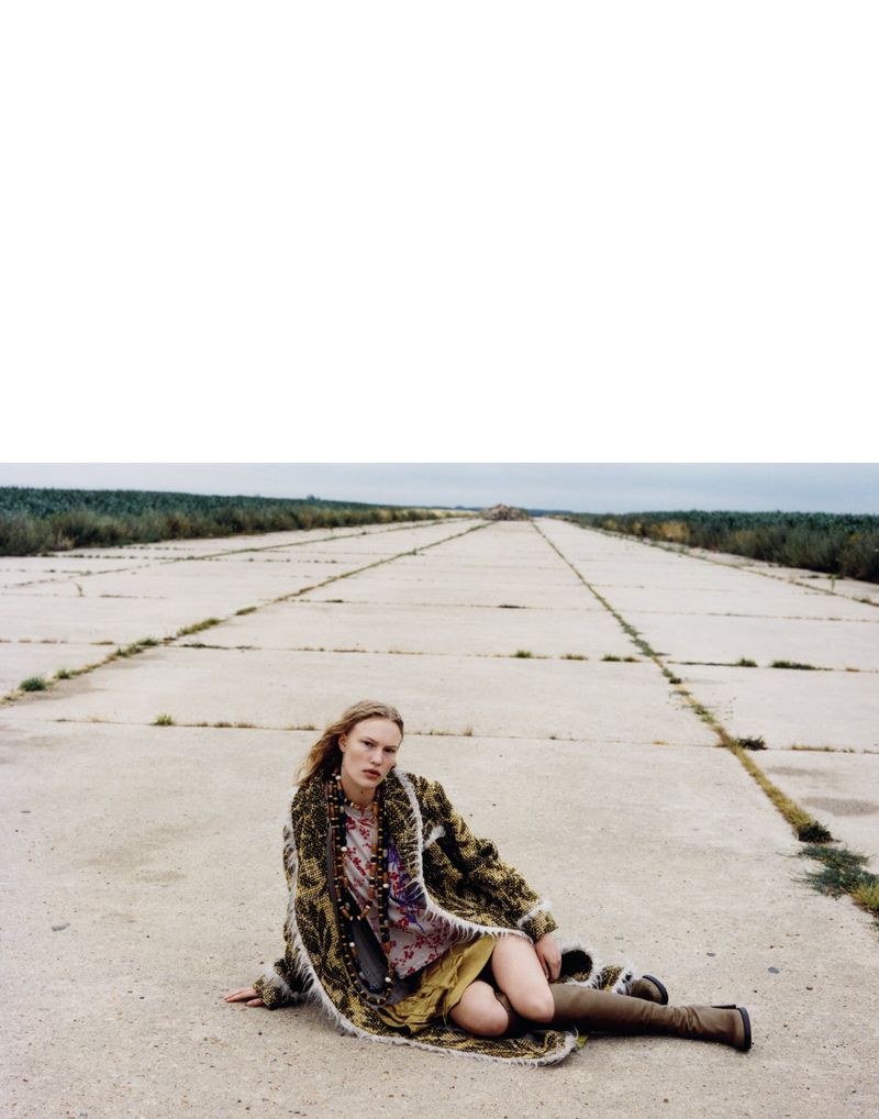 Woman in printed dress and fur coat sitting on a concrete runway outdoors on an overcast day. She is wearing high boots.
