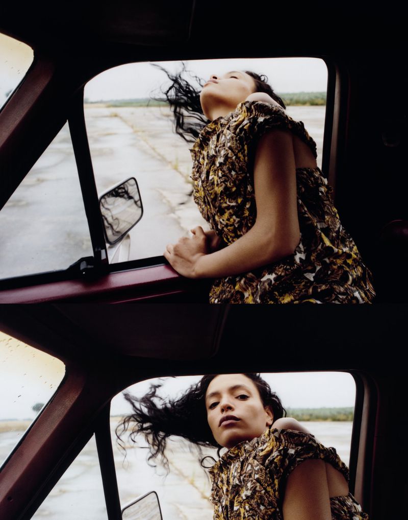 An adult female in patterned dress leans out of a car window with blowing dark hair, capturing a windy, outdoor fashion editorial shot.