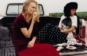 Two women sitting in the open bed of a pickup truck, one eating an apple and the other wearing a large black furry hat.