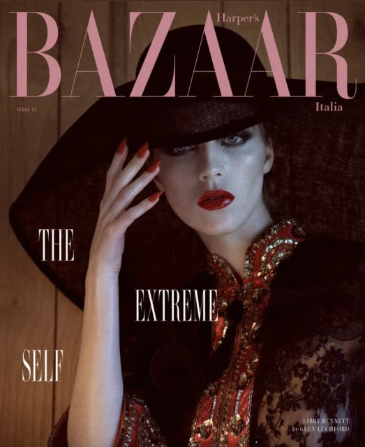 Fashion editorial cover featuring a single model in a wide-brimmed hat and decorative dress against a dark, moody backdrop.