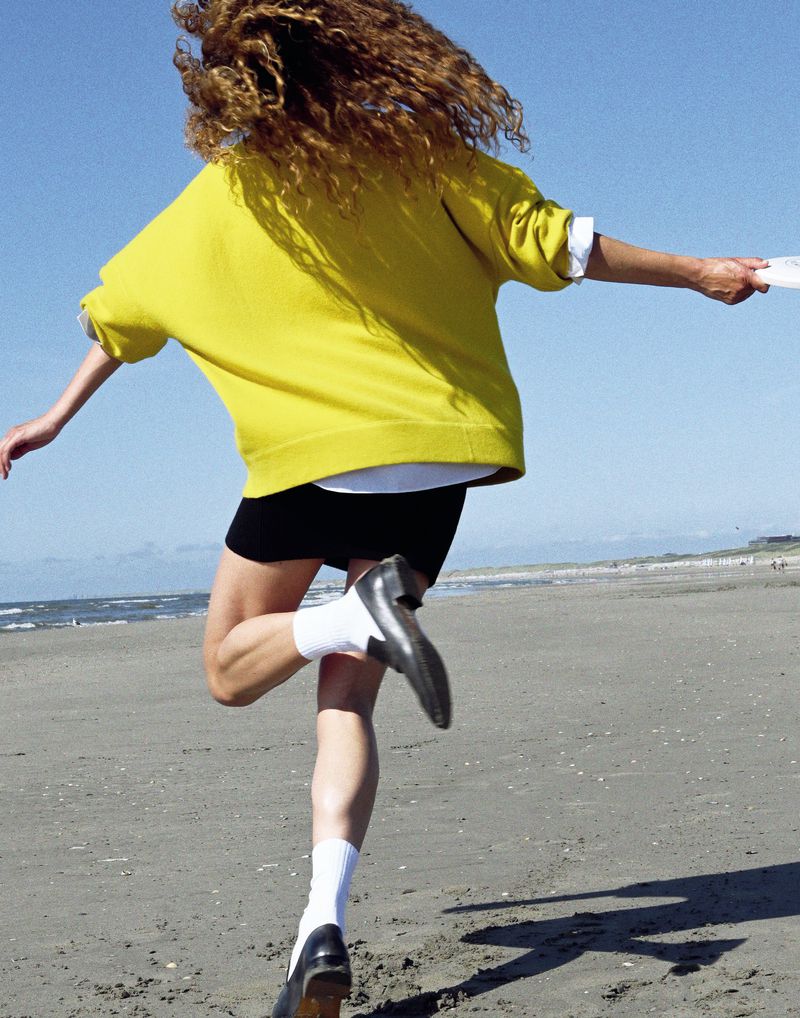 Woman running on a beach with a yellow sweater and black shorts, viewed from behind, with the ocean and blue sky in the background.