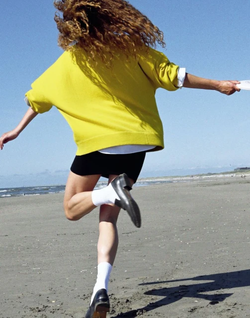 Woman running on a beach with a yellow sweater and black shorts, viewed from behind, with the ocean and blue sky in the background.