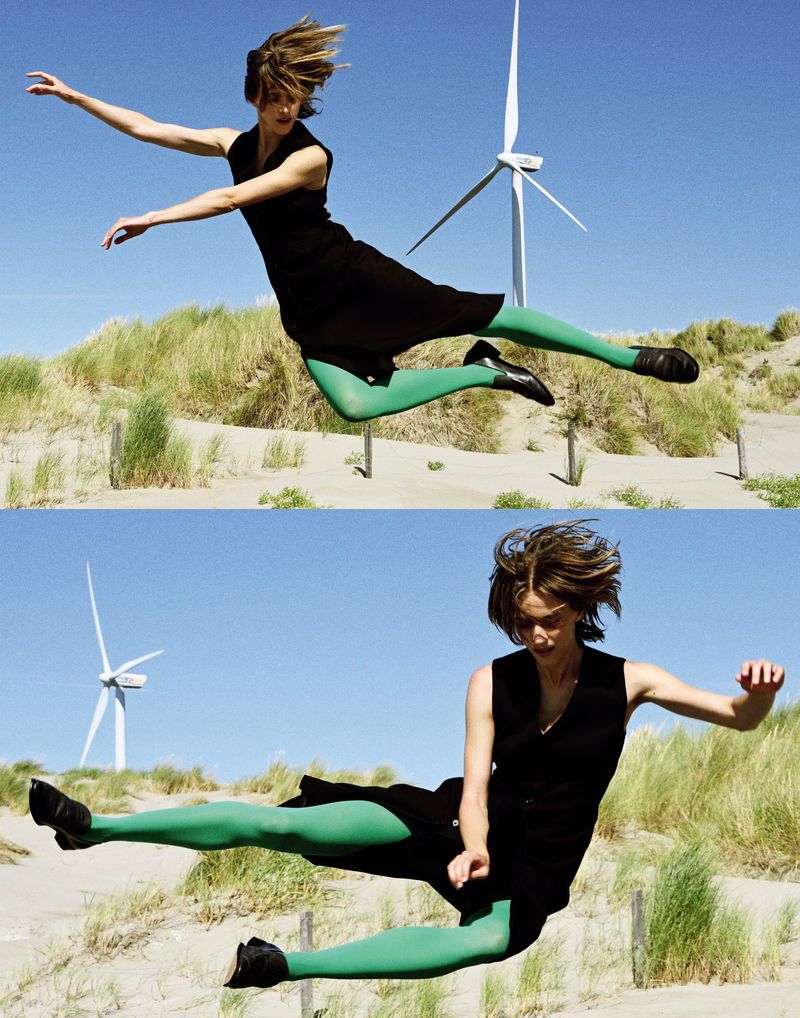 A woman in a black dress and green tights leaps on a sand dune with a wind turbine in the background on a clear, sunny day.