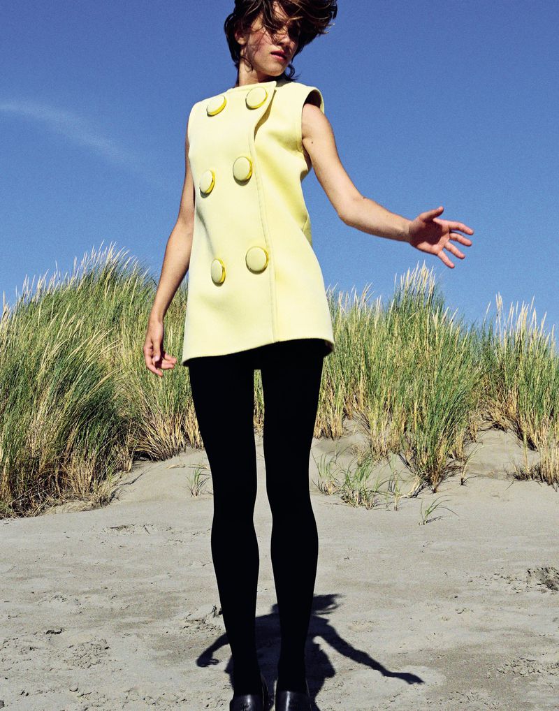 A young, slender female model poses on a sandy beach wearing a pale yellow sleeveless dress, black tights and black shoes against a blue sky.