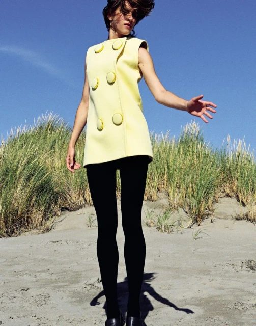 A young, slender female model poses on a sandy beach wearing a pale yellow sleeveless dress, black tights and black shoes against a blue sky.