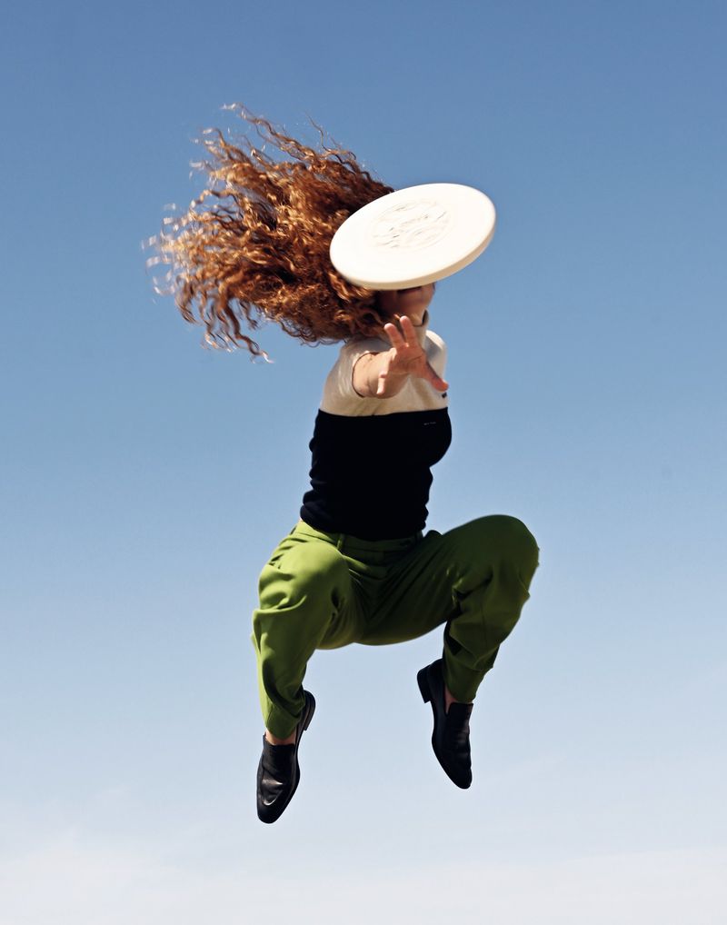 A woman with curly red hair is jumping mid-air in front of a blue sky while reaching for a white frisbee during daytime.