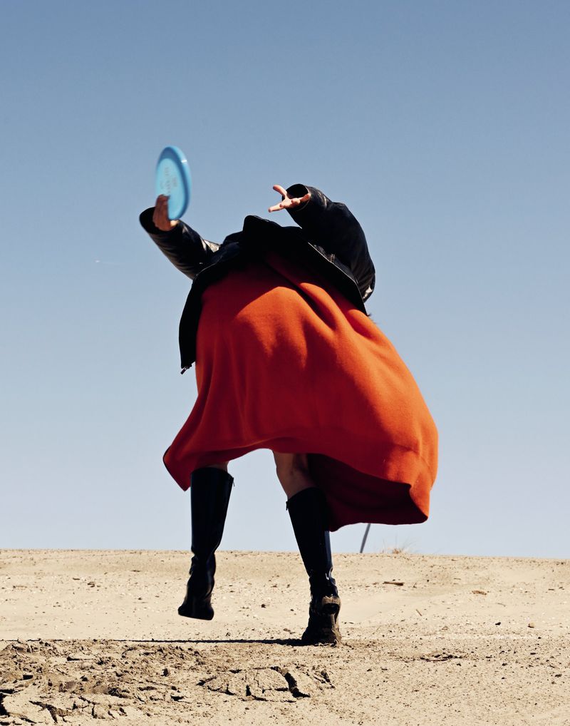 Person in black jacket and orange dress throws blue disc in the air while jumping in a sandy outdoor environment with a clear sky.