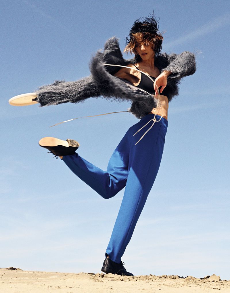 Woman in avant-garde fashion strikes dynamic pose against clear blue sky. Wearing a gray faux fur jacket, blue pants, and black boots.