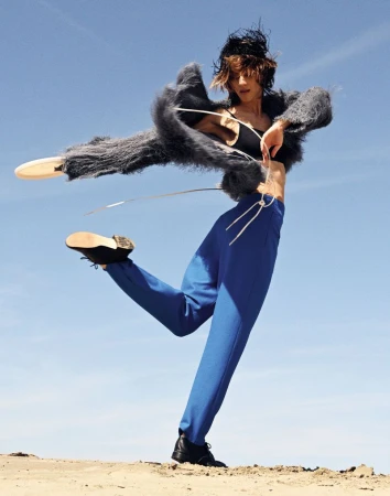 Woman in avant-garde fashion strikes dynamic pose against clear blue sky. Wearing a gray faux fur jacket, blue pants, and black boots.