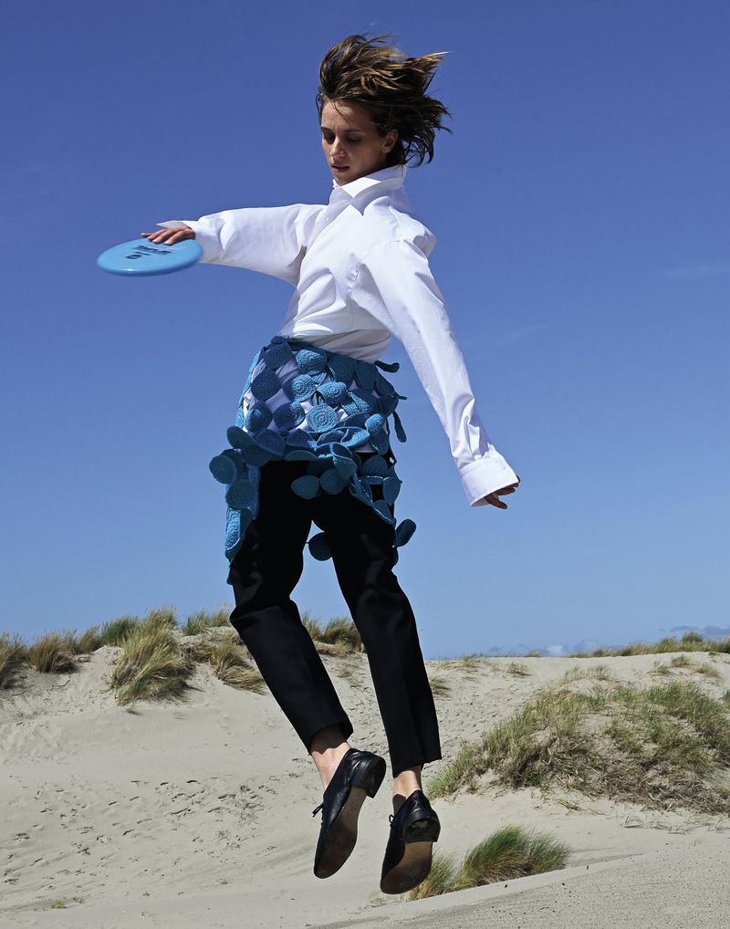 A young adult female jumps on a sandy beach, holding a blue disc against a clear blue sky. She wears a white collared shirt and black pants.