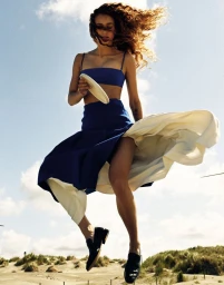 Model in blue two-piece outfit with billowing skirt mid-air against coastal dunes and blue sky for Annabelle Magazine.