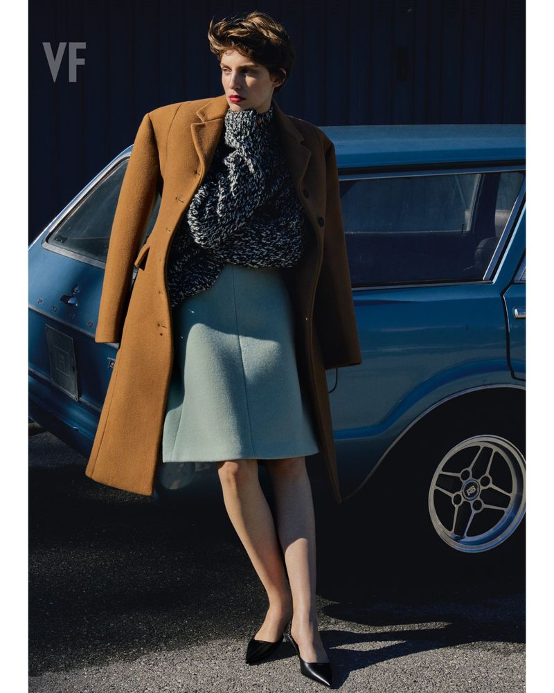 Elegant woman in camel coat, textured sweater, and mint skirt posing by a classic light blue car on an asphalt street.