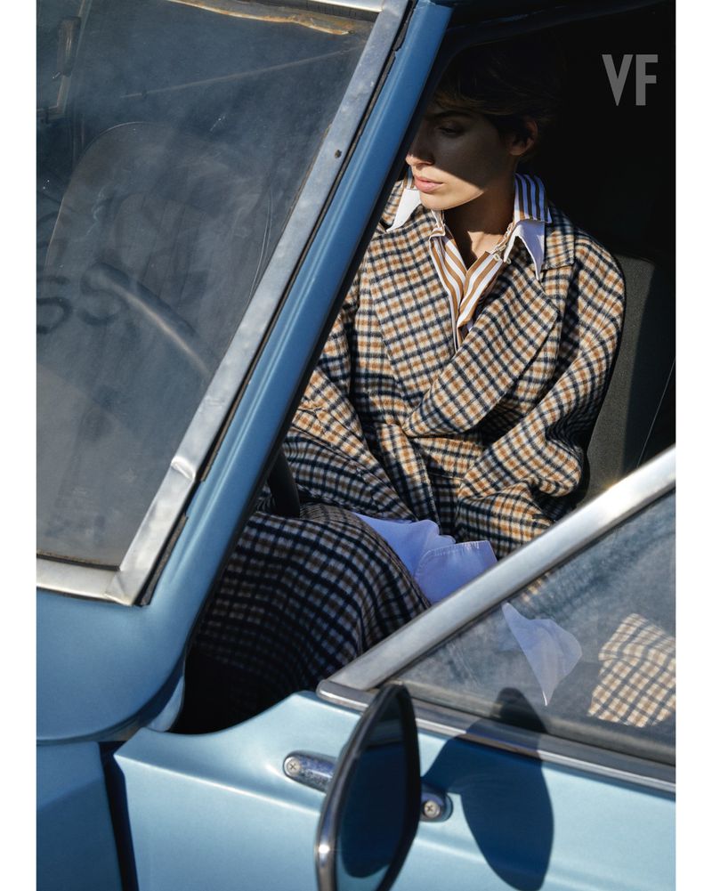 Woman in plaid coat sits inside light blue vintage car, looking down. Soft light. Vanity Fair logo upper right. Automotive editorial.