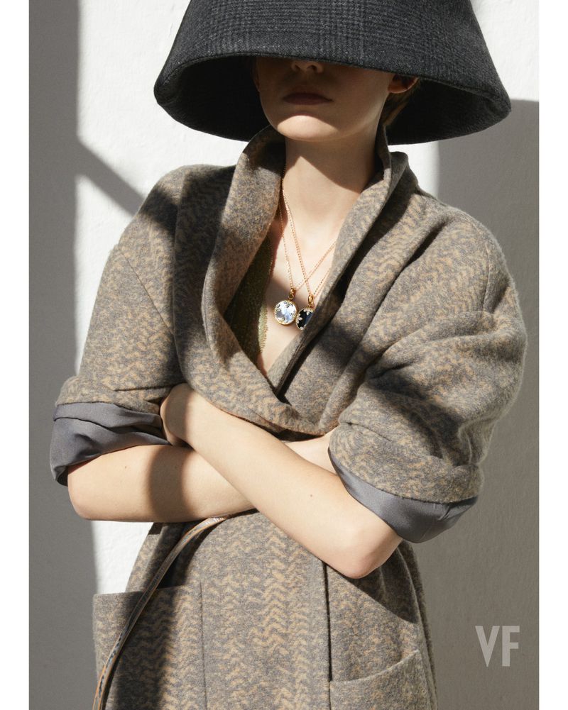 Fashion shot of a woman wearing a gray patterned coat and a dark gray wide-brimmed hat, accessorized with gold necklaces and pendants.