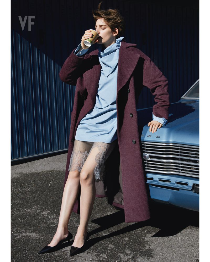 Woman in a stylish coat and dress holds coffee and leans against a vintage car. Blue wall behind.