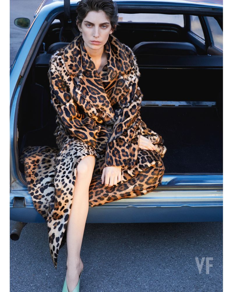 Woman in a leopard-print coat sits in the open trunk of a car, looking at the camera. Fashion editorial with vintage auto.