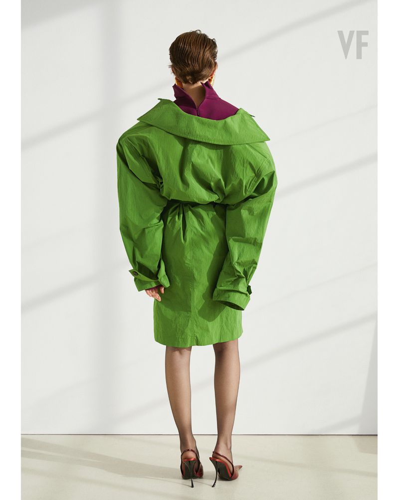 Back view of a female fashion model in a green oversized dress with long sleeves and red high heels against a white background.