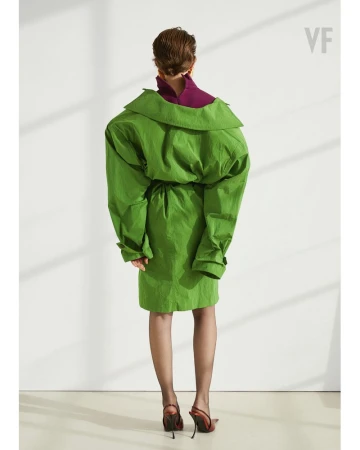 Back view of a female fashion model in a green oversized dress with long sleeves and red high heels against a white background.