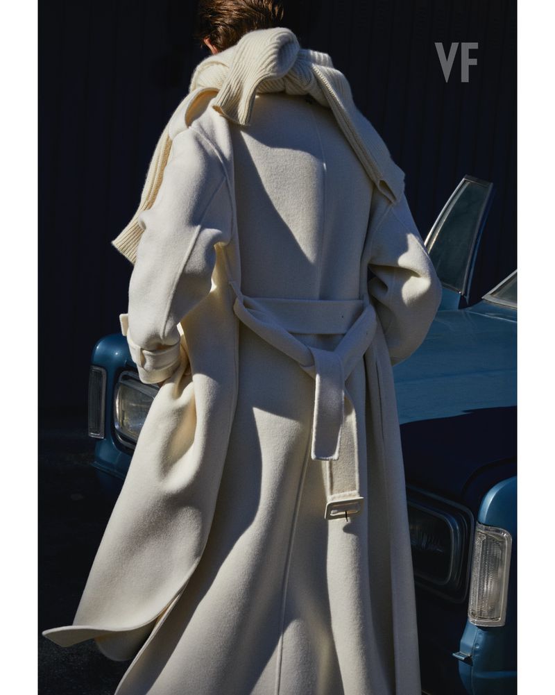 Person from behind wearing a belted white coat with a matching scarf, standing next to a blue classic car in soft, diffused light.