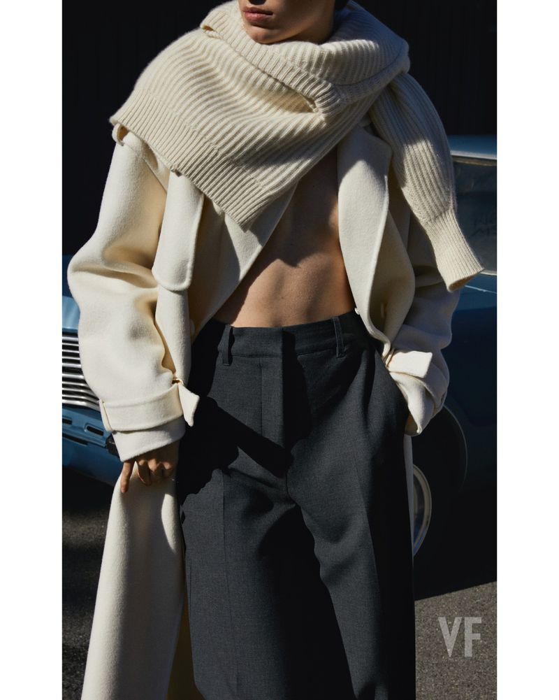 Fashion editorial shot of a stylish woman wearing a cream coat and scarf, paired with grey pants, in front of a vintage car.