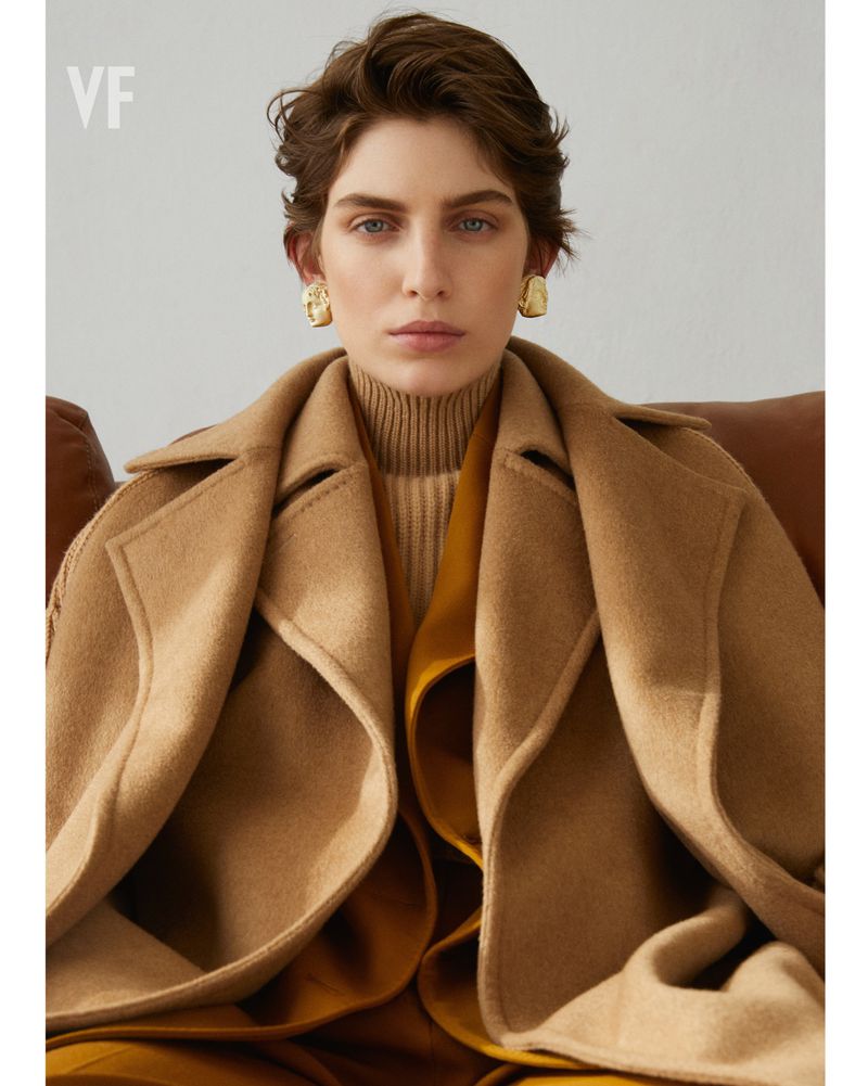 Brunette adult female in a tan overcoat and gold earrings, posing indoors in front of a light neutral background, fashion portrait.