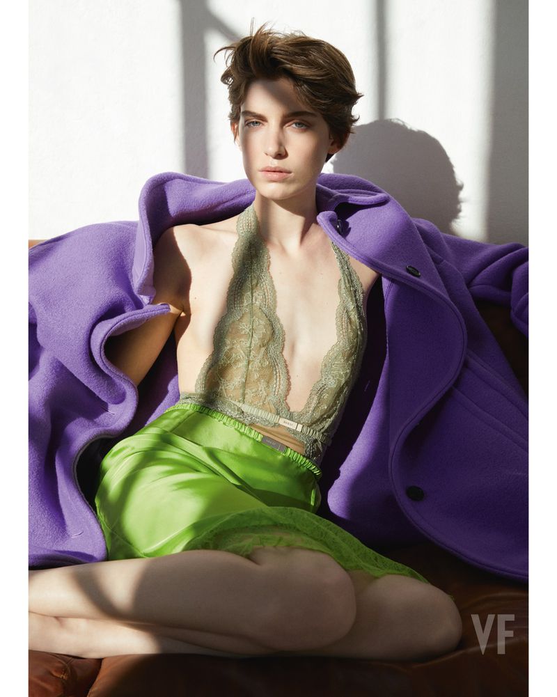 Fashion photo of a woman with short hair sitting and wearing a purple coat, a lace top, and a green skirt, indoors with shadows.