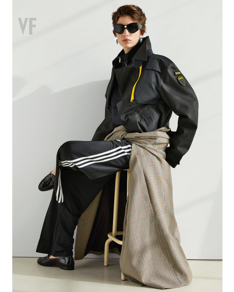 A person wearing a black trench coat and sunglasses sits on a stool, showcasing fashion against a plain background in a studio setting.