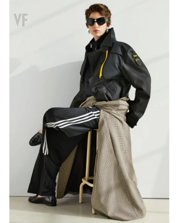 A person wearing a black trench coat and sunglasses sits on a stool, showcasing fashion against a plain background in a studio setting.
