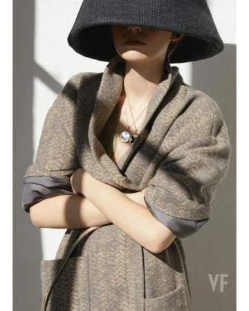 Model wearing a wide-brimmed black hat and patterned coat with layered gold necklaces against a light wall casting diagonal shadows